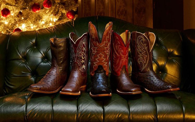 A row of Tony Lama boots on a green leather couch with a glowing Christmas tree in the background.